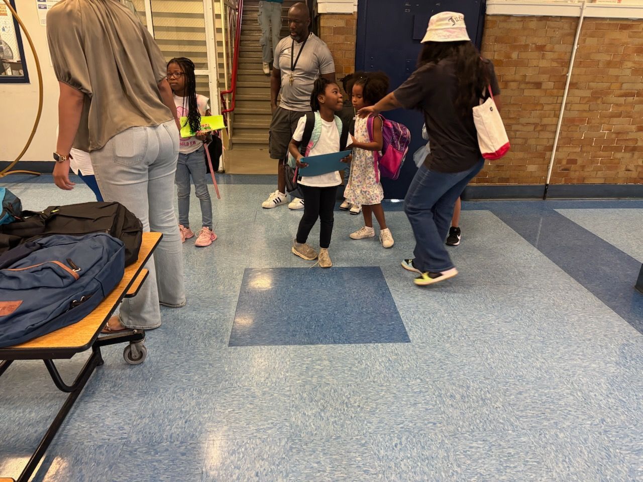 People in a hallway, some wearing backpacks. One person dances. Blue and light-colored floors.