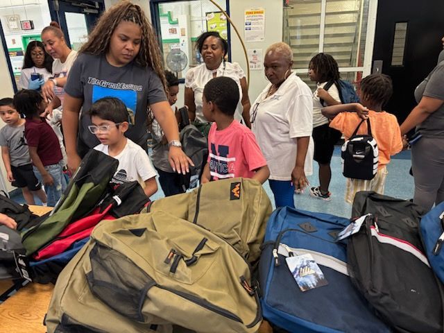 People selecting from a pile of backpacks. Inside, adults and children. Some touch the bags, appearing ready to begin school.