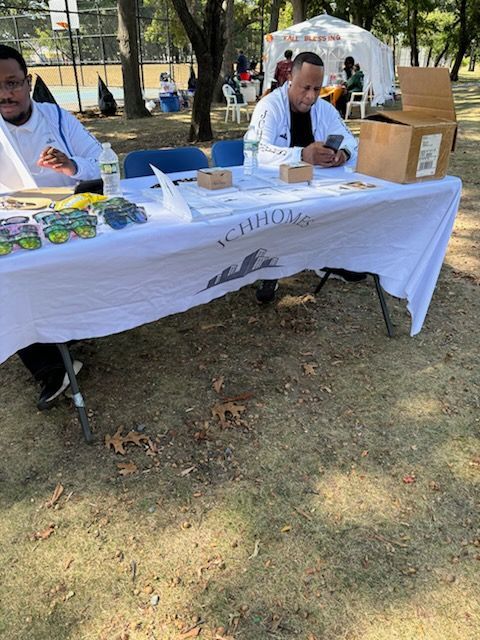 Two men at a table with merchandise and a sign in a park setting. One man on phone.