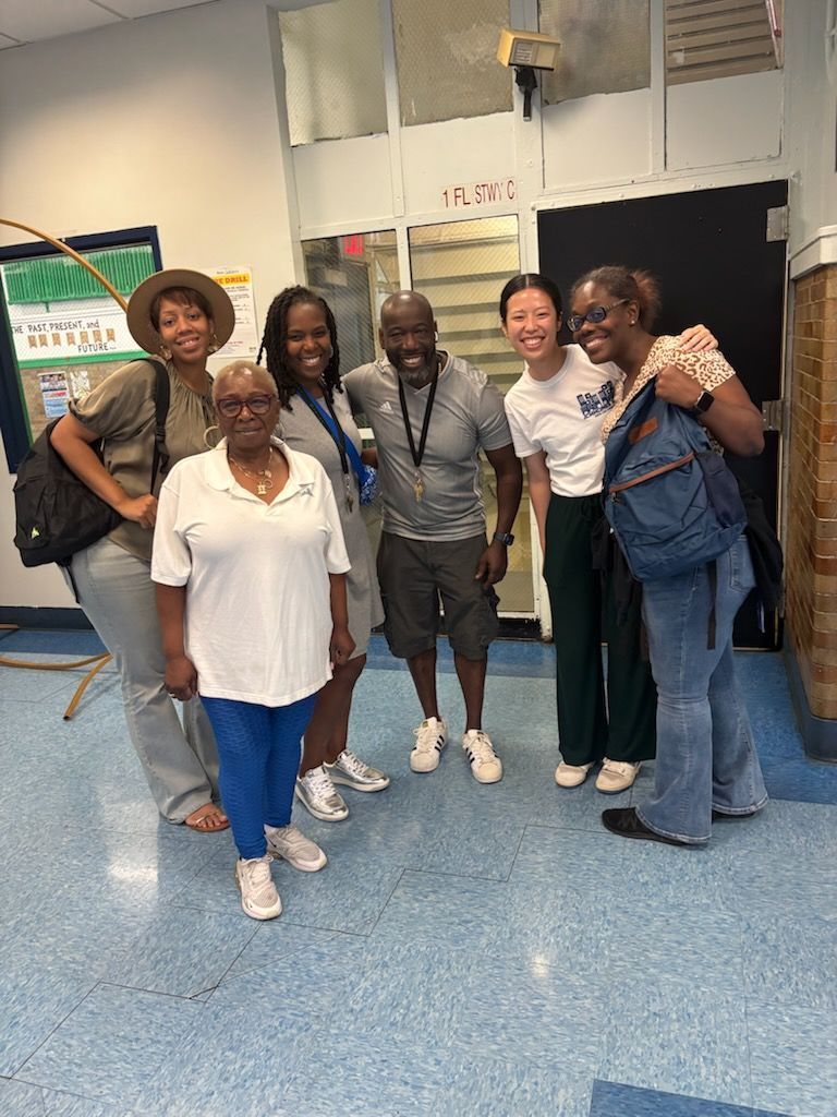 Group of people smiling, standing in a hallway. One person is wearing a hat, another a backpack, and one with arms around others.
