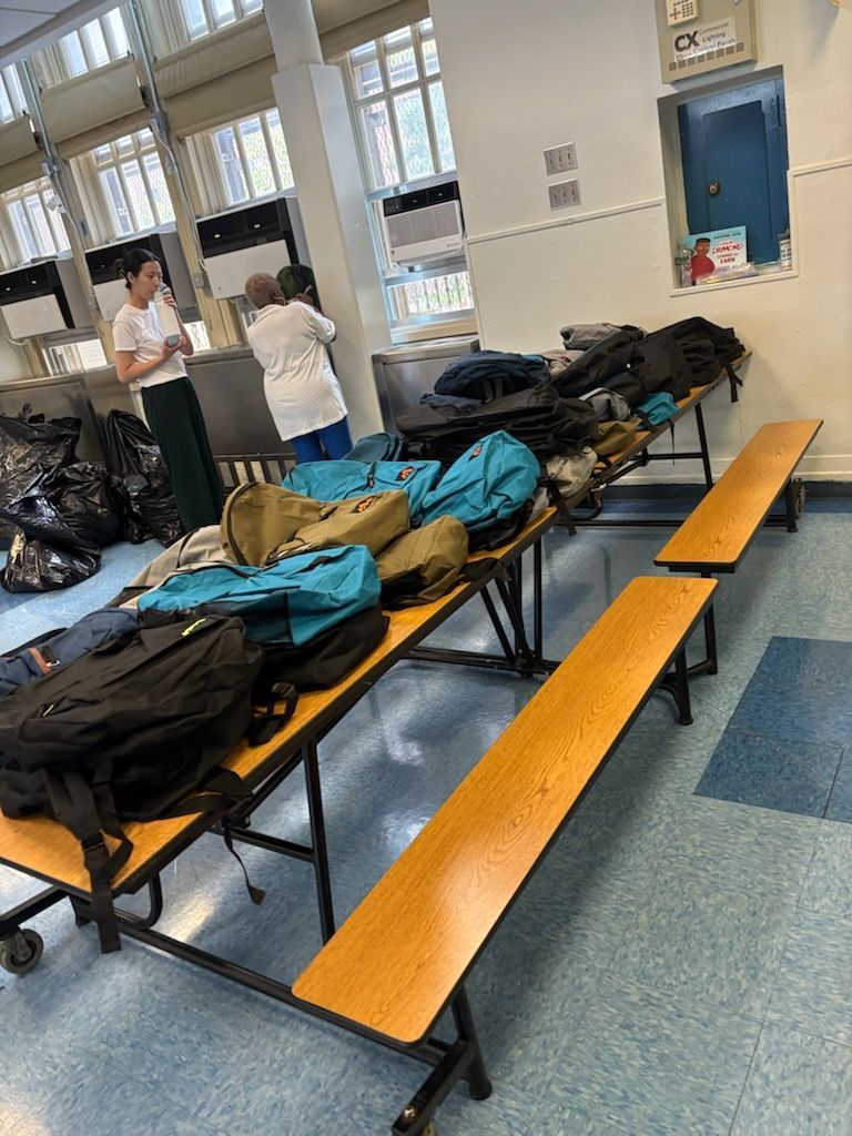 Lunchroom: Backpacks on tables, two women near a counter, and benches. Blue floor and white walls.