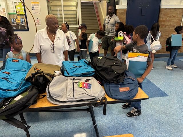 People selecting backpacks from a table. Several kids and an adult are present indoors. Various colors of backpacks are displayed.