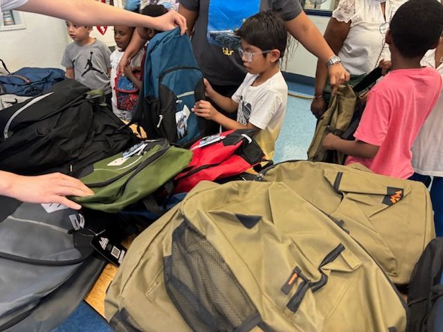 Children picking out backpacks at a donation event. Various colors and styles of backpacks on a table.