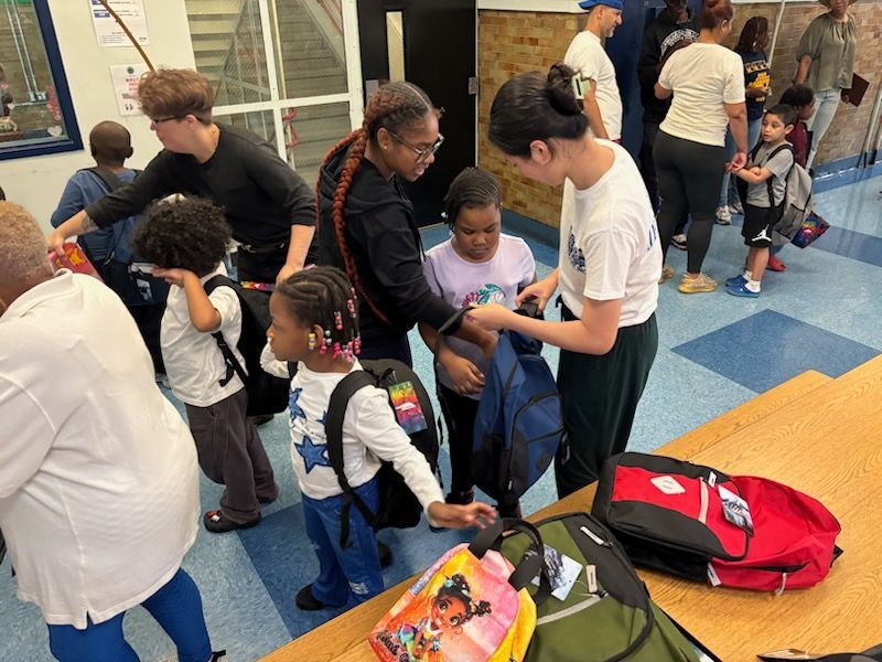 People distributing backpacks to children in a brightly lit room.
