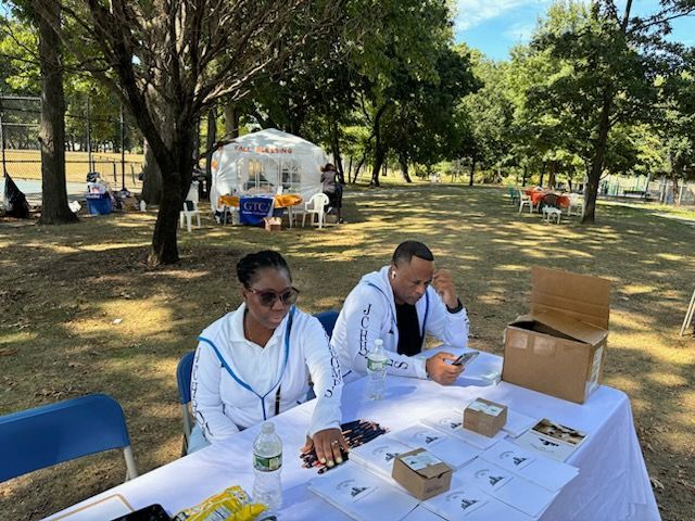 Two people at a park table with papers and boxes, a tent in the background.