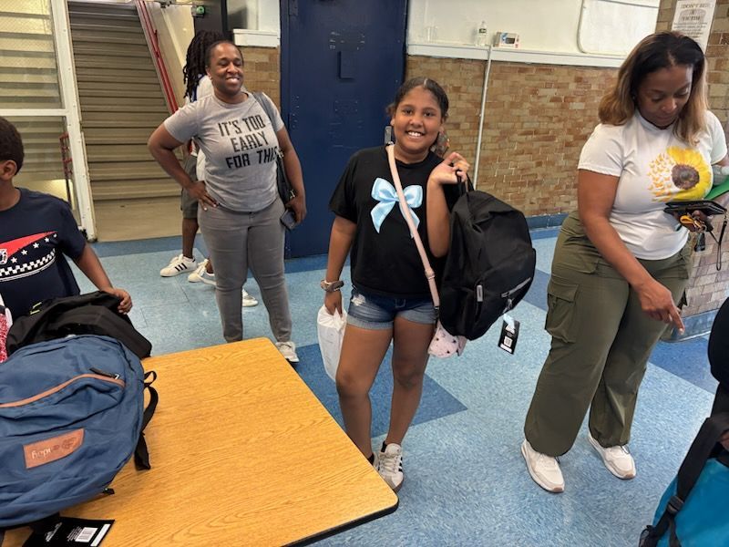 Girl with backpack smiles, surrounded by adults and other backpacks.