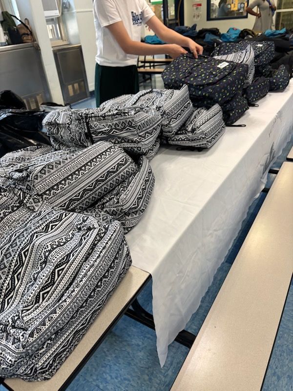 Person arranging many patterned backpacks on a white-covered table. Indoors, likely a school or event.