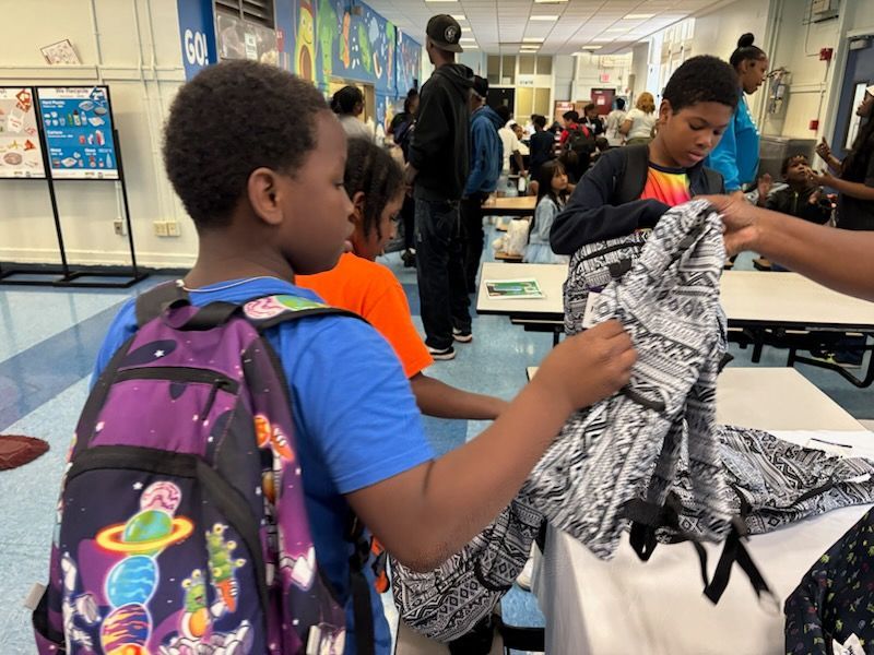 Kids looking at clothing in a school cafeteria. Tables hold clothes for them to choose from. Others watch.