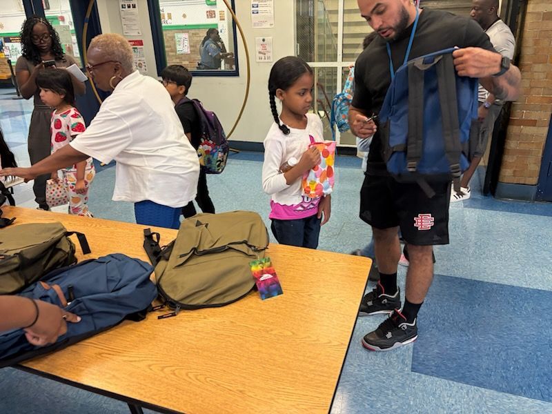 People at a table distributing backpacks. A child looks on, holding a book, in a school setting.