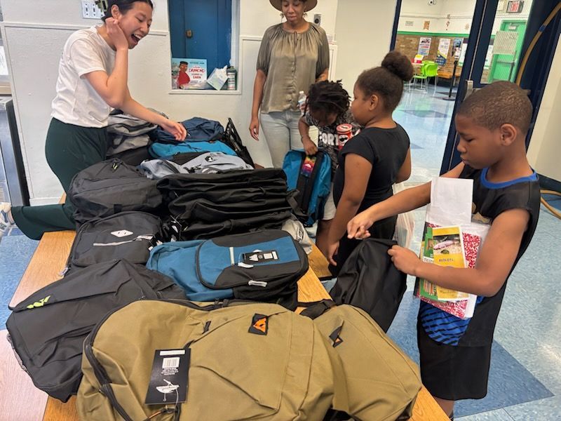 People and children selecting backpacks from a table, indoors.
