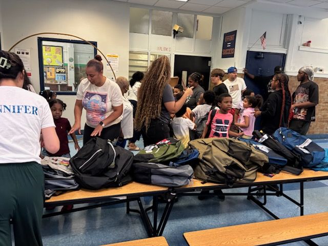 People gather around tables piled with backpacks indoors; adults assist children.
