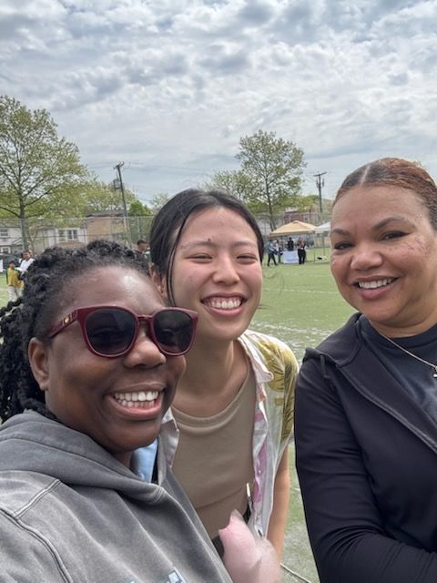 Three smiling people pose for a selfie outdoors on a cloudy day, field in background.