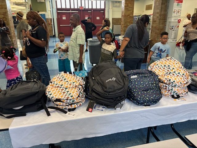 People gather near a table displaying backpacks. Children and adults appear to be selecting them. Indoors, brightly lit.