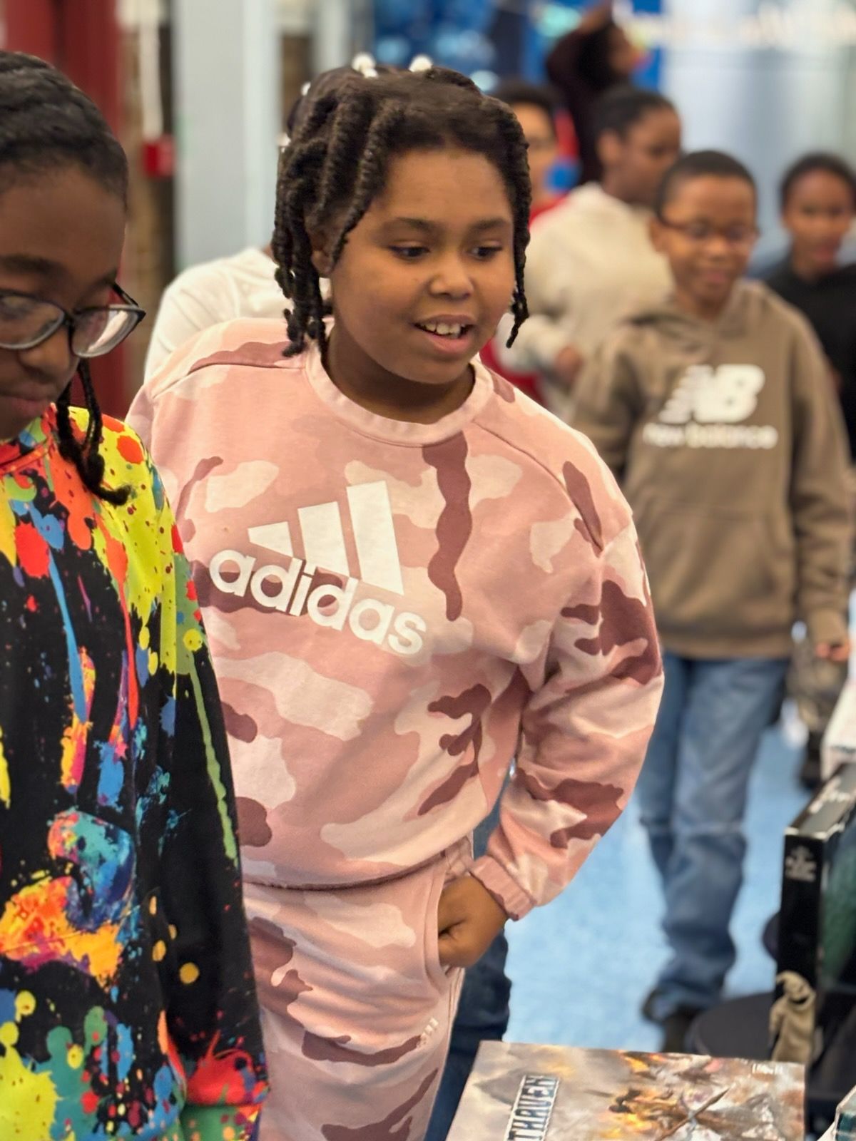 Girl in pink camo Adidas sweatshirt looks at a table with others in a brightly lit room.