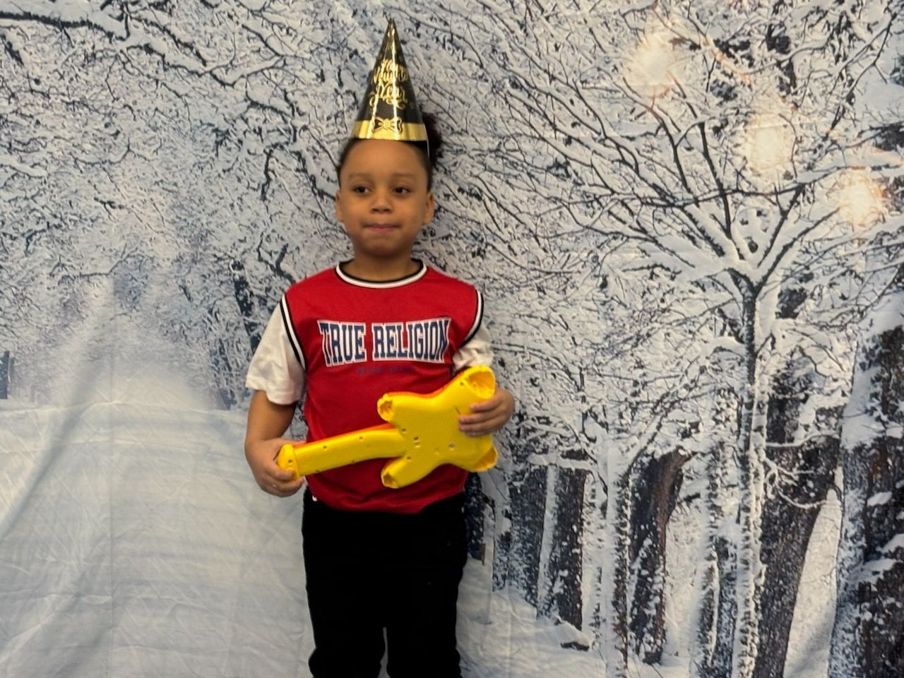Child in party hat and red shirt holding a yellow star, standing in front of a winter backdrop.