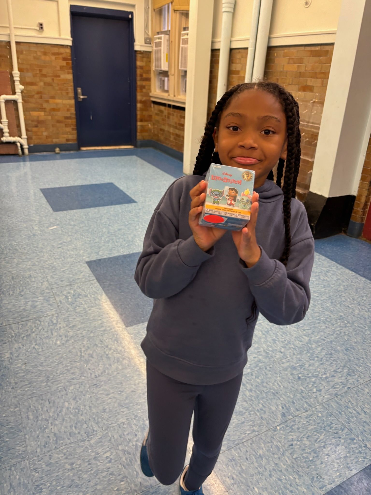 Girl in blue sweatsuit holding a box with both hands, smiling, inside a school hallway.