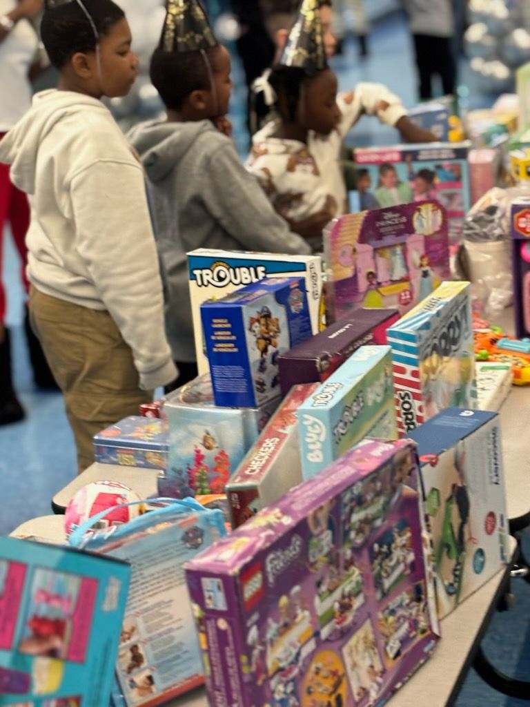 Children wearing party hats look at a table laden with wrapped gifts, including toys and games.