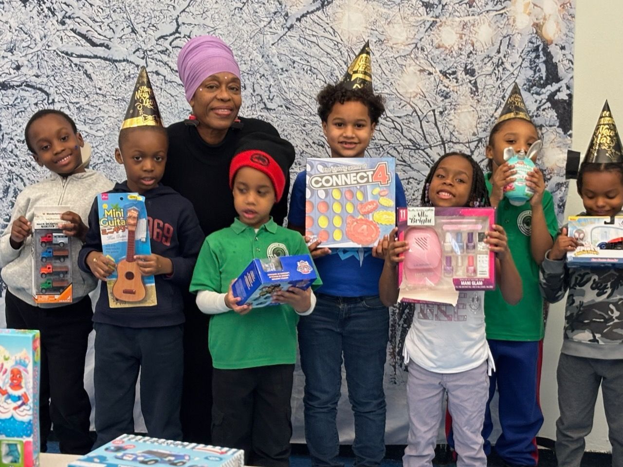 Group of children and an adult with party hats, holding gifts, in front of a colorful backdrop.