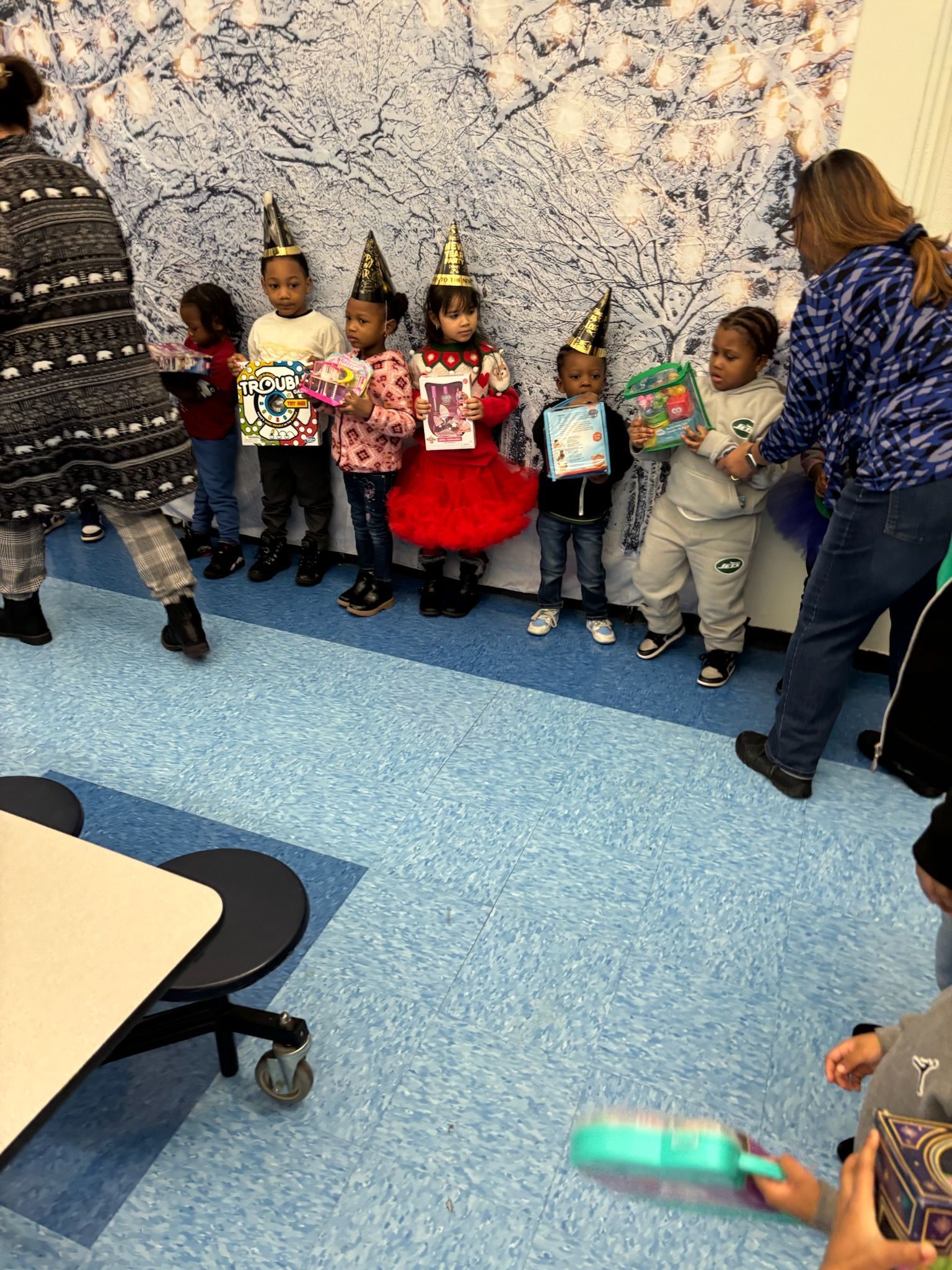 Children with party hats, holding items, stand in a line. Adults oversee them. Blue and white patterned wall.