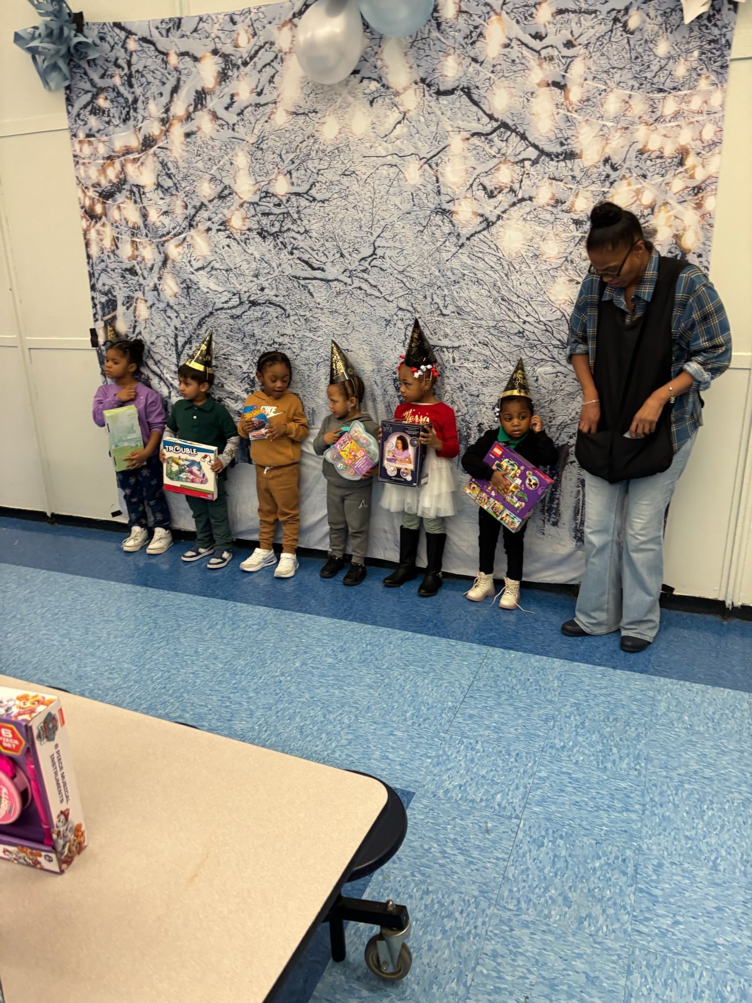 Children in party hats stand in front of a winter-themed backdrop, holding books. An adult stands to the right.