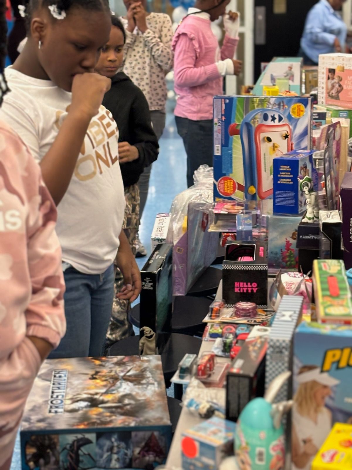 Children browsing toys at a table; focus on a girl looking thoughtful. Variety of brightly colored toys visible.