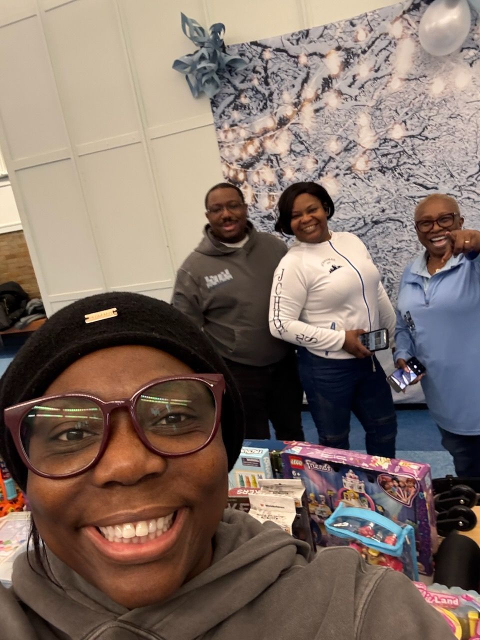 Four people smiling, posing with toys in front of a blue and white backdrop. One person takes a selfie.