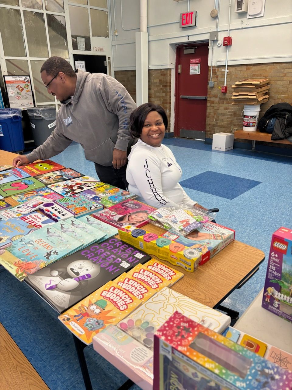 Two people at a table displaying books and gifts, smiling. Indoor setting with a blue floor.