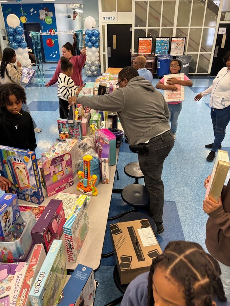 People at a table selecting gifts at an event, many toys are displayed. Balloons and other decorations in a school setting.