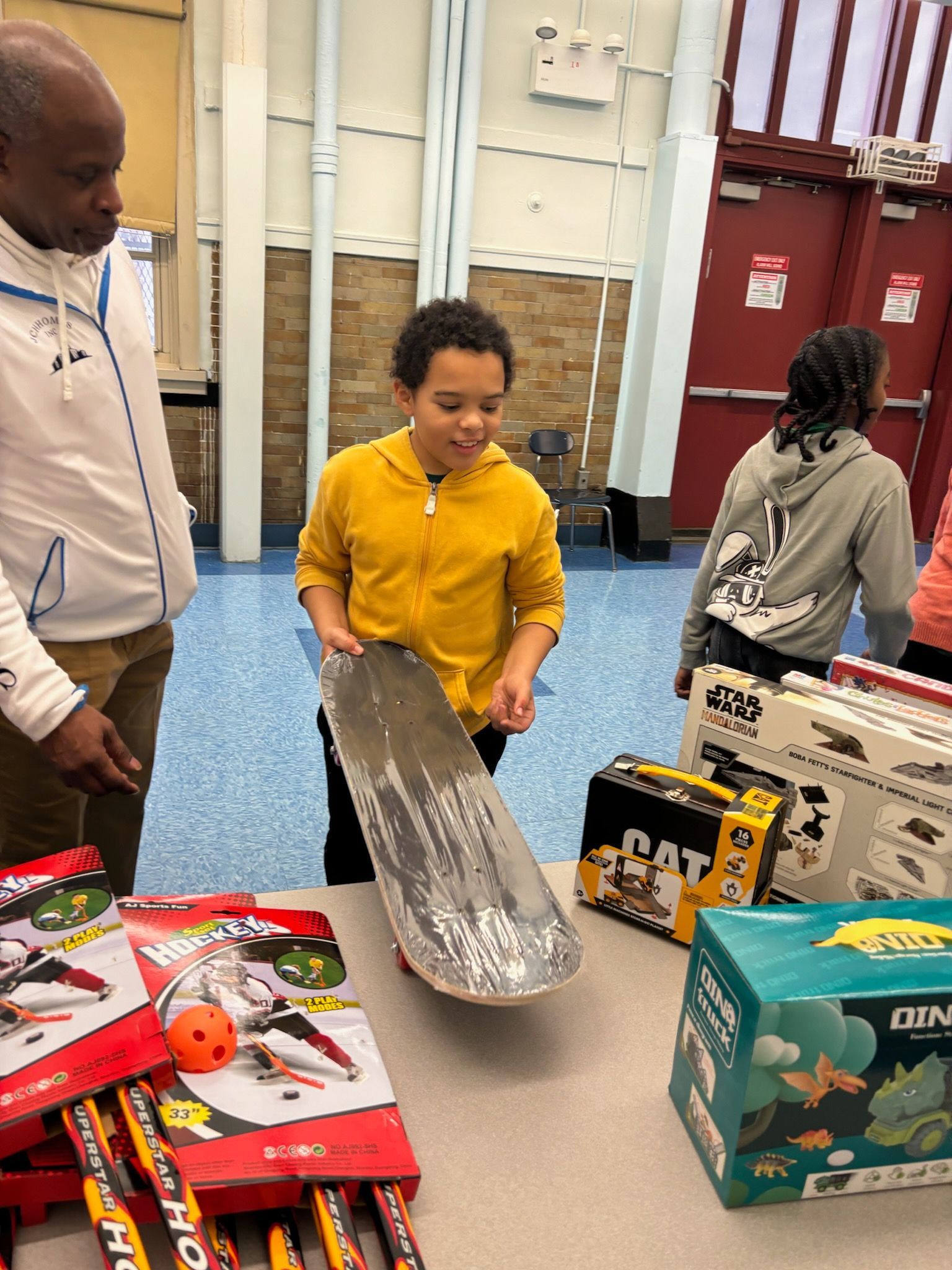 Boy holding a skateboard, looking at it, with a man watching. Toys on a table.