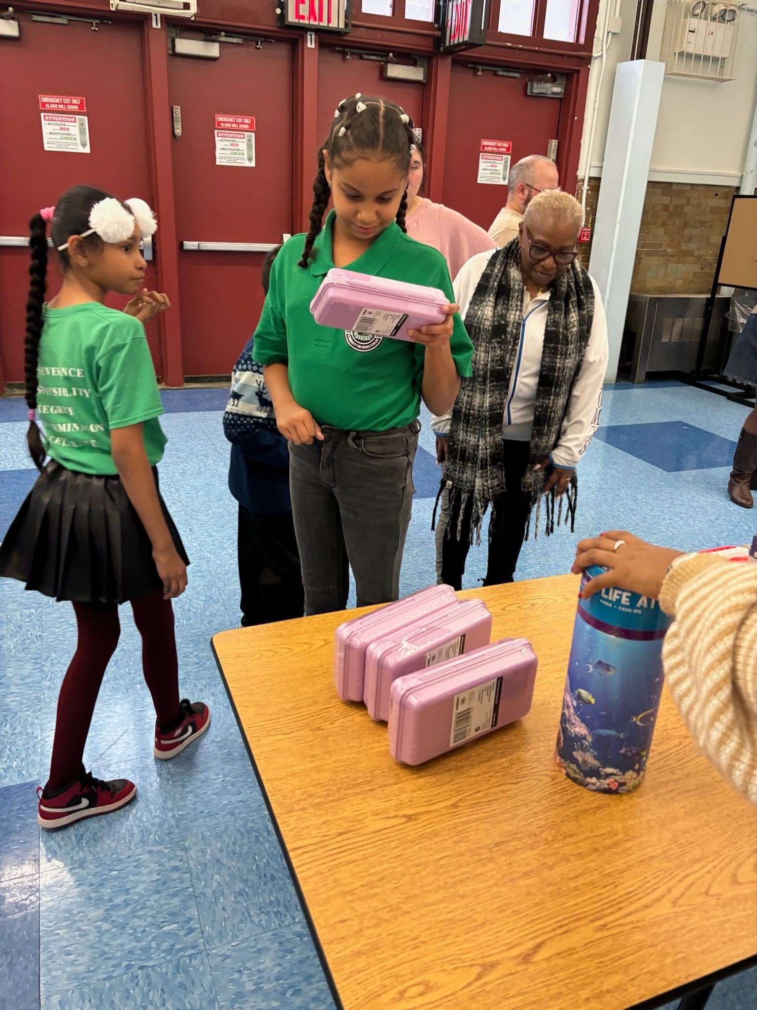 Two girls and an adult look at stacked pink boxes on a table, another person beside them. School hallway.