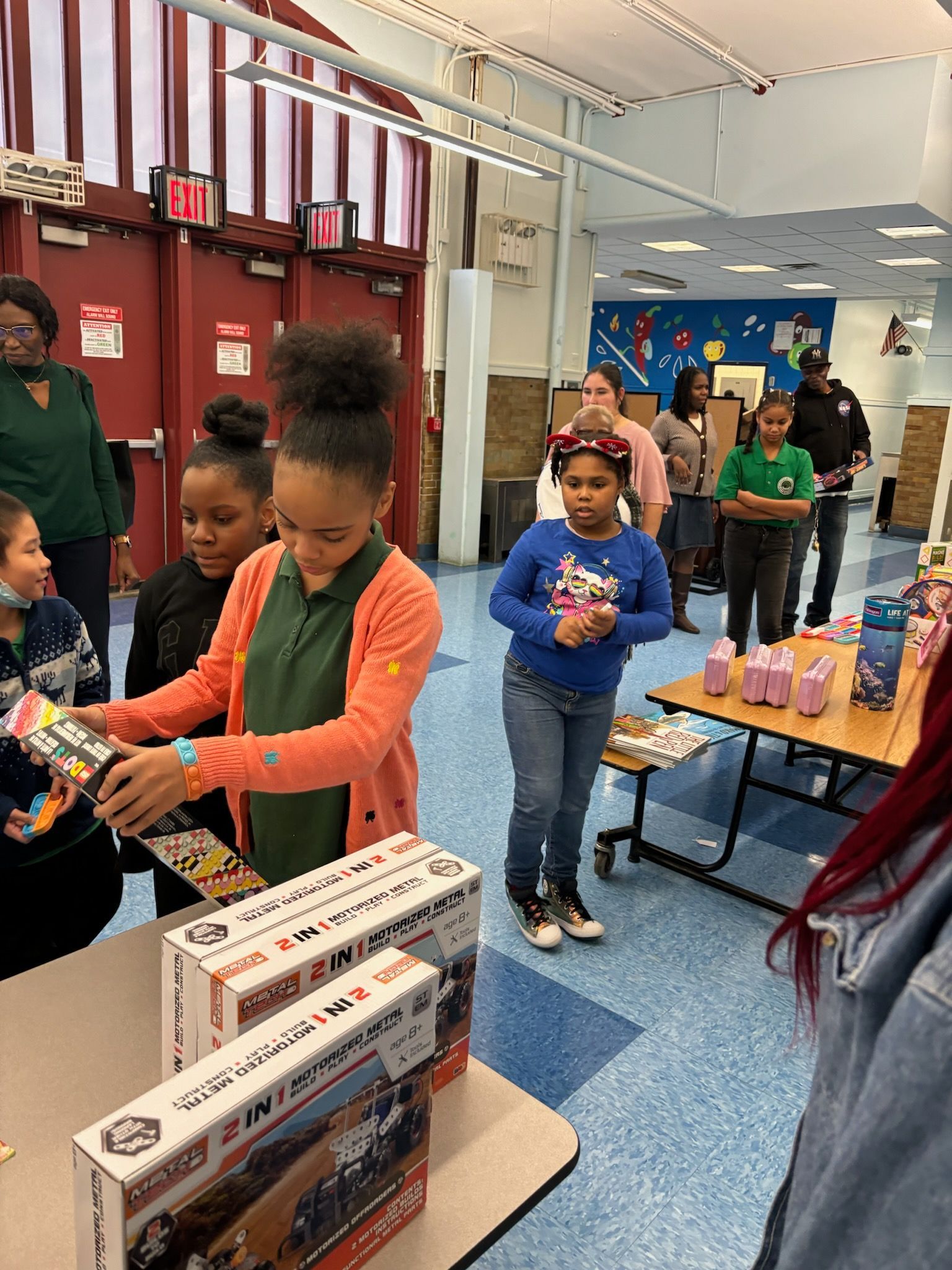 Children at a table, receiving gifts, other people in the background, likely an event inside a building.