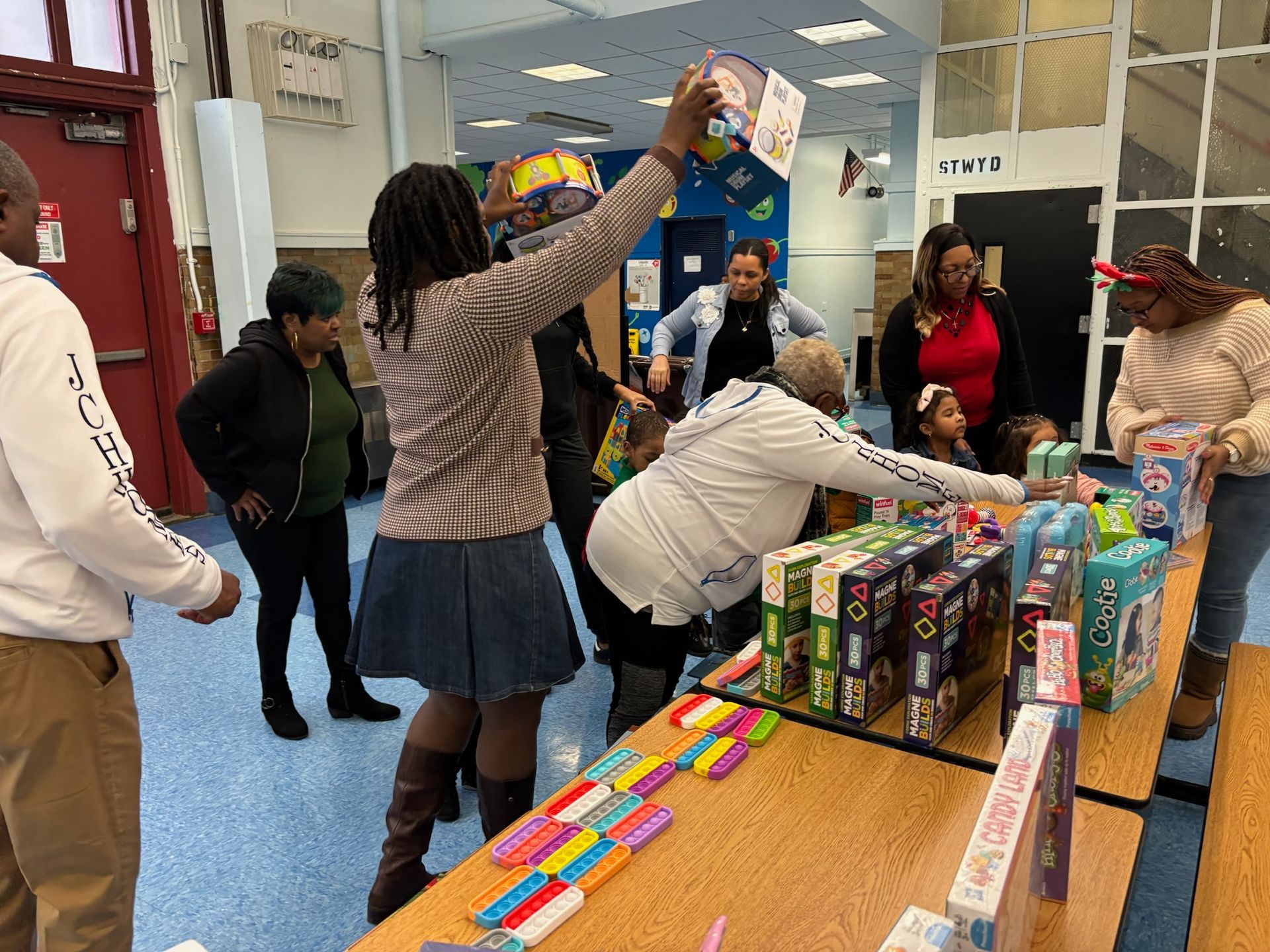 People at a table with toys, one person lifting a box.  Inside a brightly lit room.