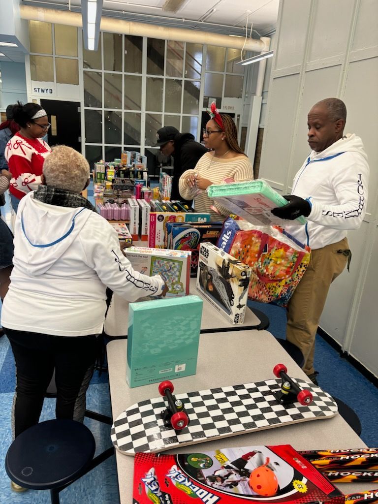 People shopping at a table laden with gifts. A person holds a skateboard. Toys and books are visible.