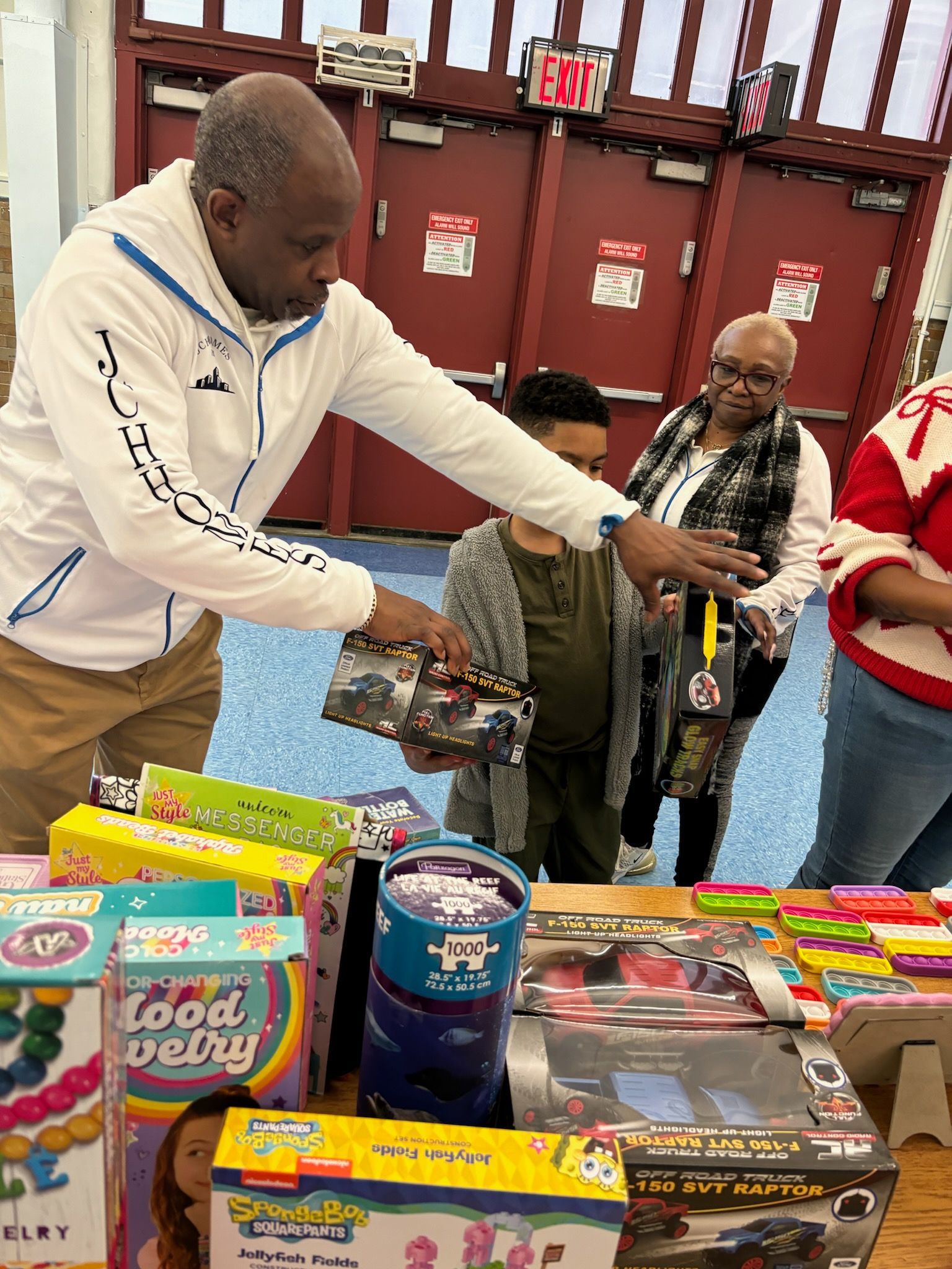 Man handing a toy to a child at a table of toys. An older person is watching. Indoors, festive.