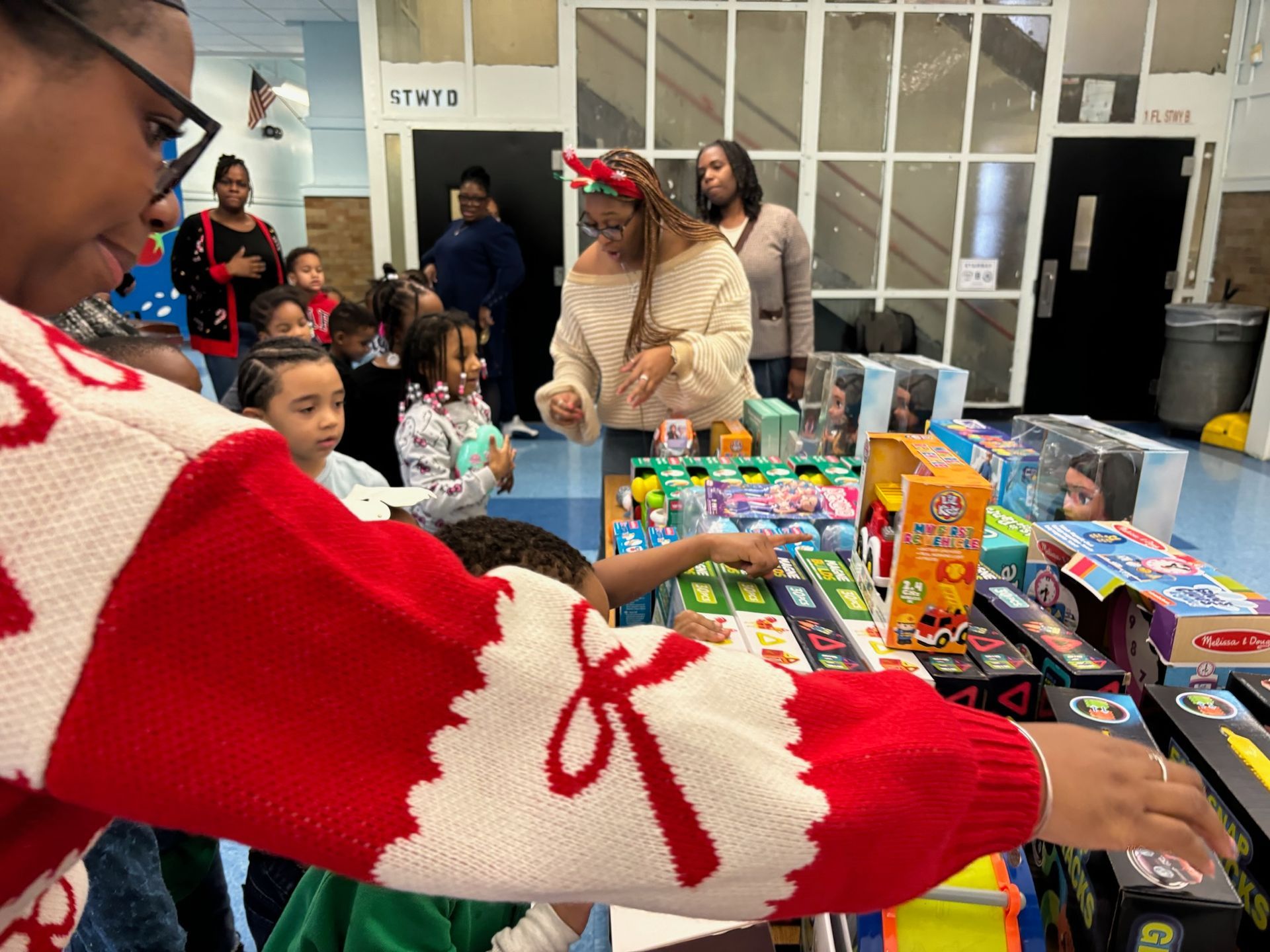 Children picking out gifts at a table, with adults looking on, in a brightly lit room.