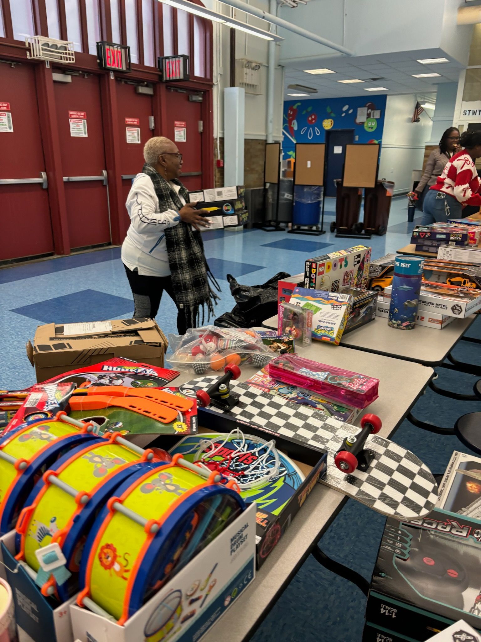 Woman with gifts at table, drums, skateboards, and other toys displayed. Interior, school event.