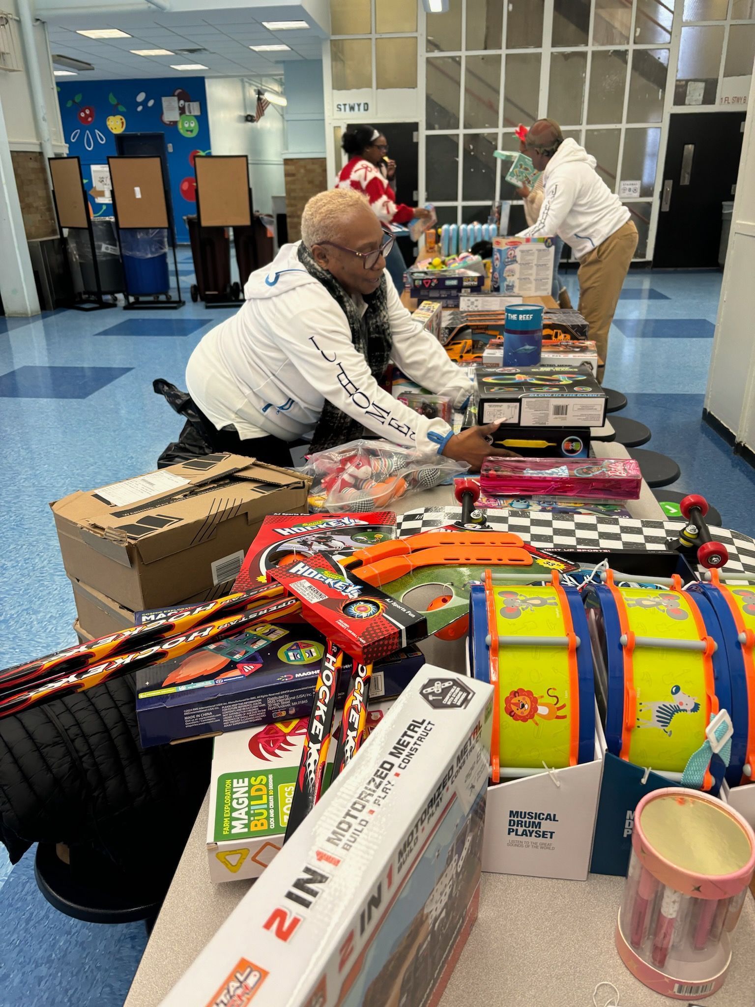 People arranging toys on a table in a brightly lit room. Boxes and containers filled with toys.