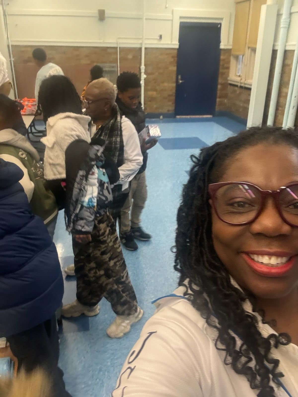 Woman in glasses smiles, taking a selfie. People in line, indoors.