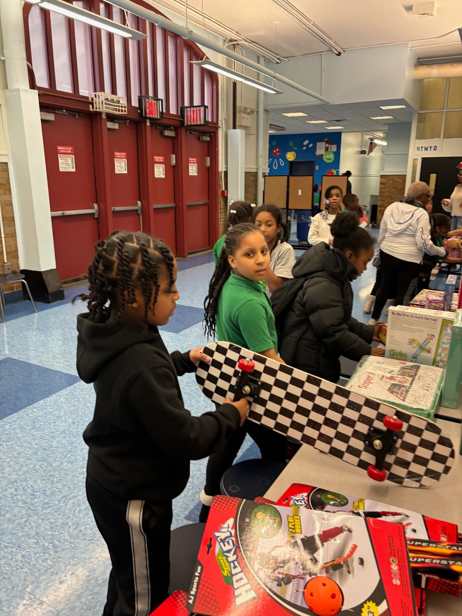 Boy holds a checkered skateboard at a school event. Other children and adults are at tables.