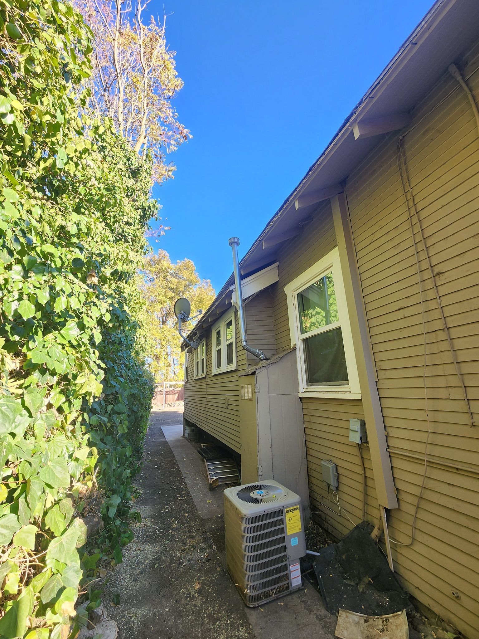 Side view of a yellow house with an air conditioning unit and narrow walkway next to a hedge under a blue sky.