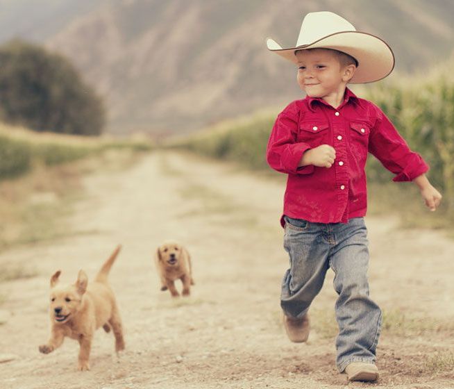 Boy in cowboy hat and red shirt running on dirt road with two puppies.