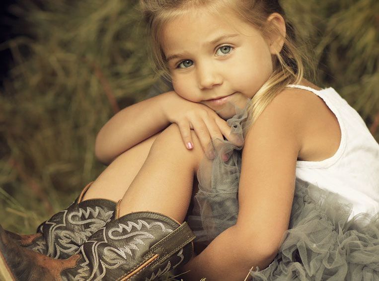 Young girl with blonde hair, blue eyes, in a white shirt and gray tutu, wearing cowboy boots, smiles, outdoor setting.