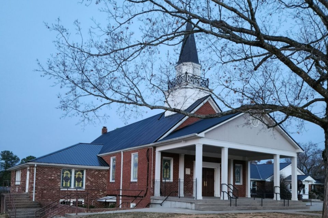 Small red-brick church with white columns and steeple under bare trees at dusk