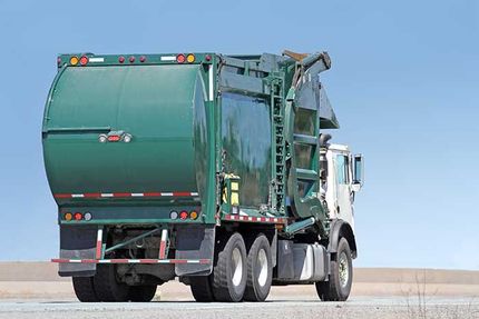 Green garbage truck on a road under a clear blue sky, viewed from the rear side.