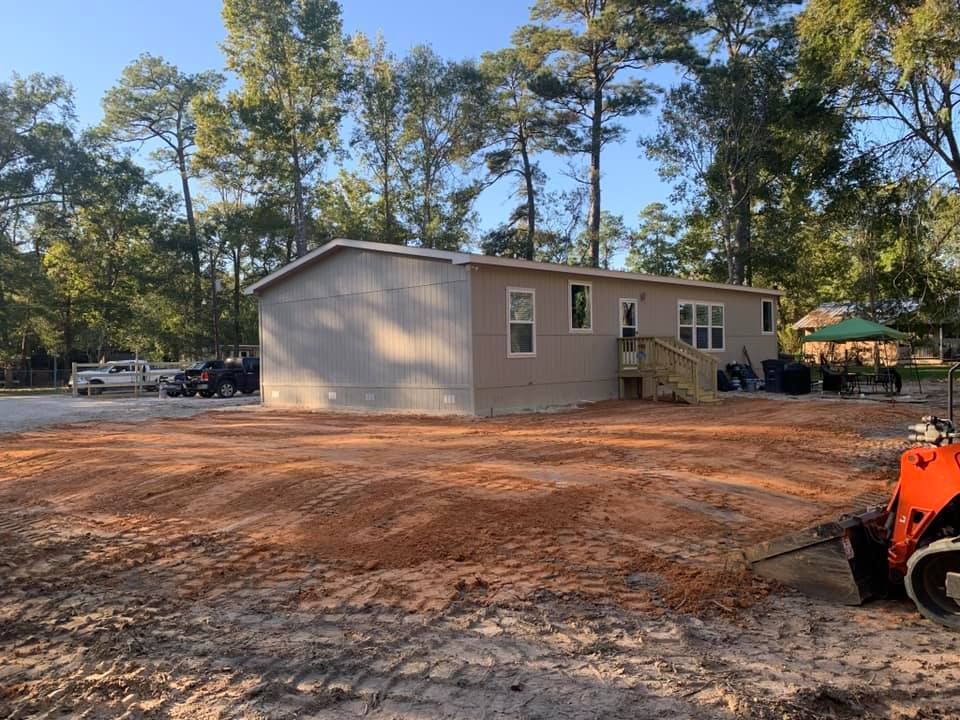 Mobile home on freshly graded dirt, surrounded by trees under a blue sky. A small construction vehicle is in the foreground.