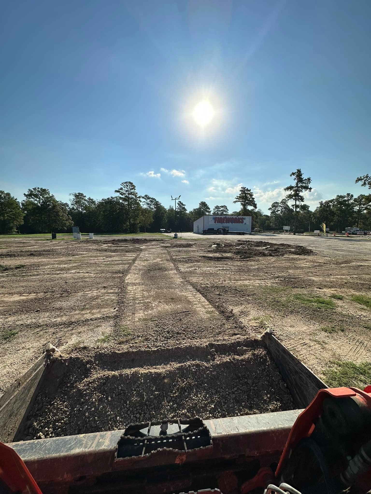 Bright sun over a gravel construction site with a white building in the background. A tractor bucket is in the foreground.