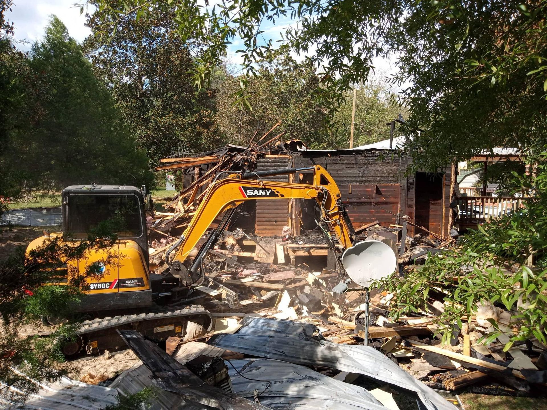 Yellow excavator demolishing a wooden structure amidst debris, surrounded by trees.