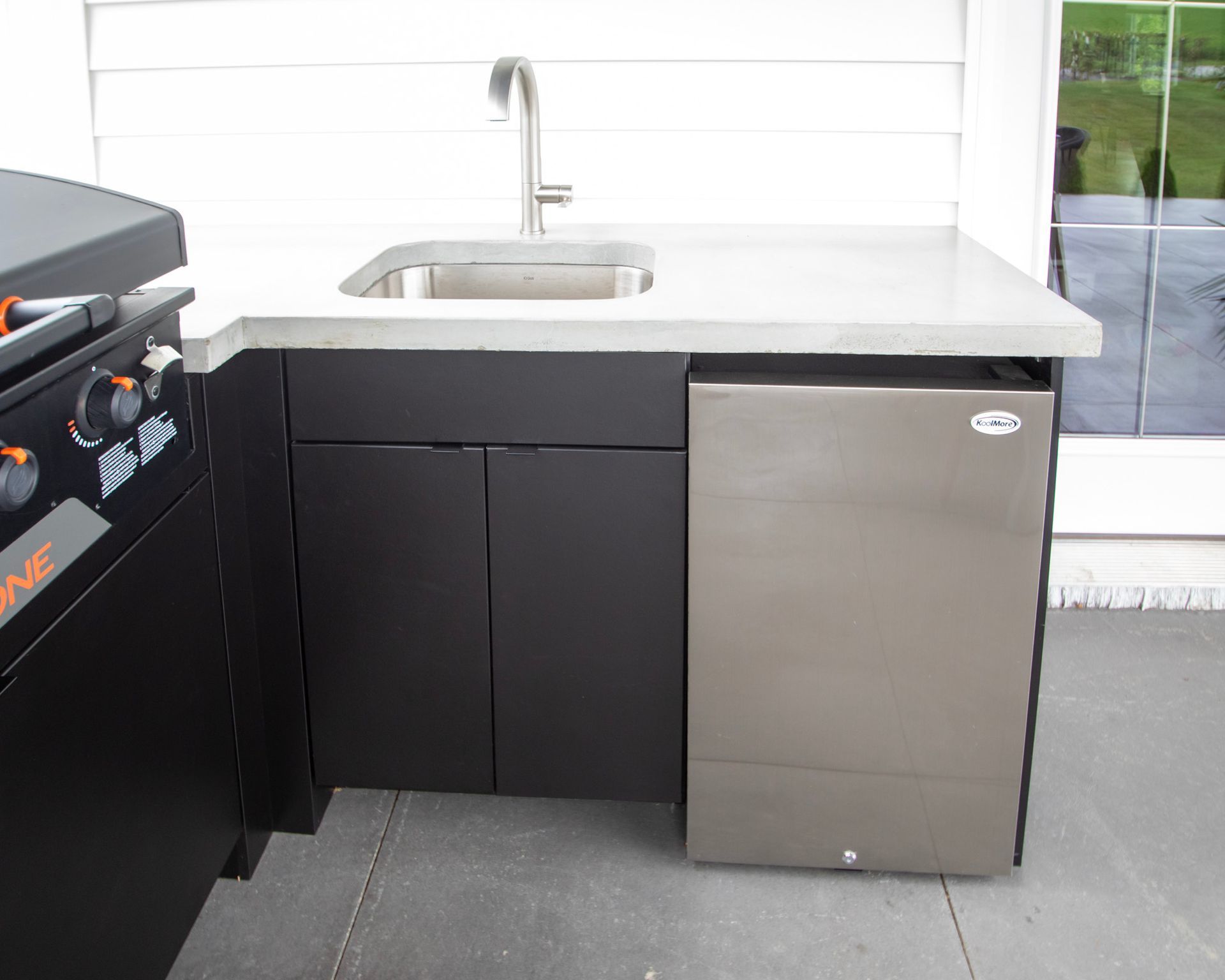 Outdoor kitchen with black cabinets, stainless sink, and mini fridge beside a grill