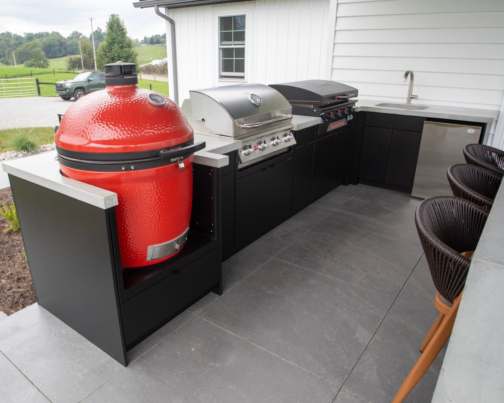 Outdoor patio with a red kamado grill, stainless grill, and black storage cabinets beside a house
