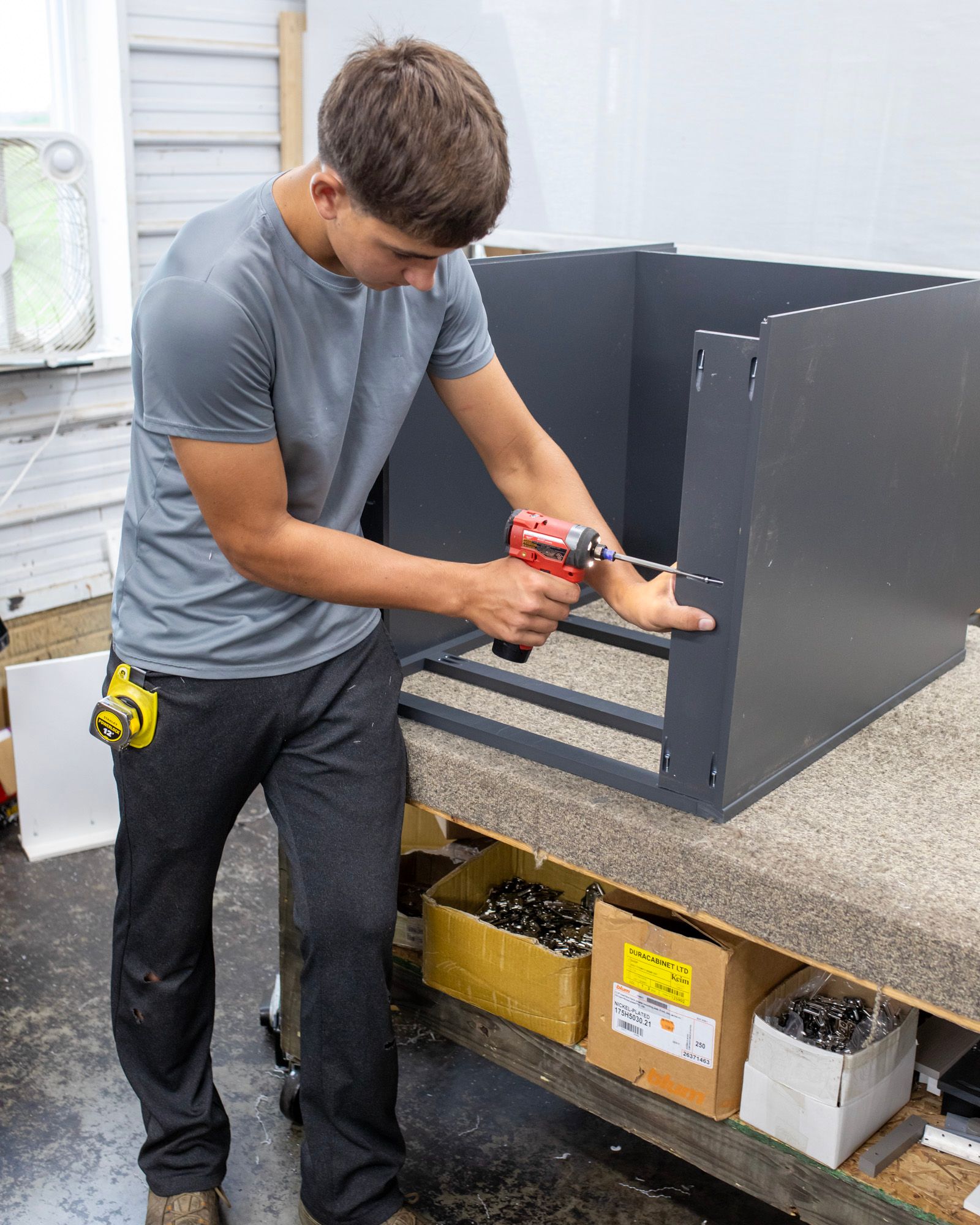 Man using a drill to assemble a gray cabinet on a workbench in a workshop